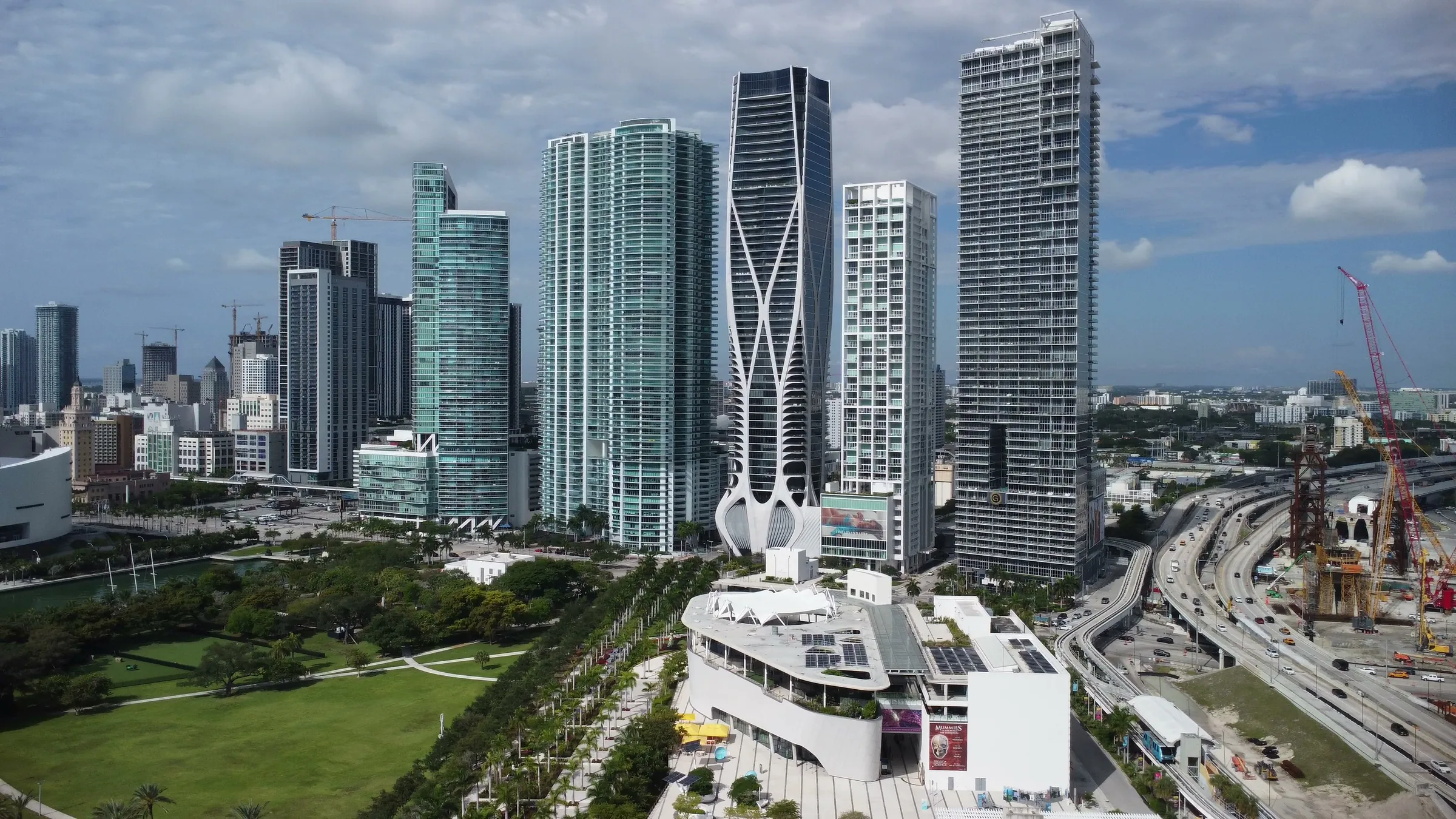Aerial view of downtown Miami: the science museum and Museum Park in front of glass residential towers, with the I-395 highway reconstruction and cranes on the right.