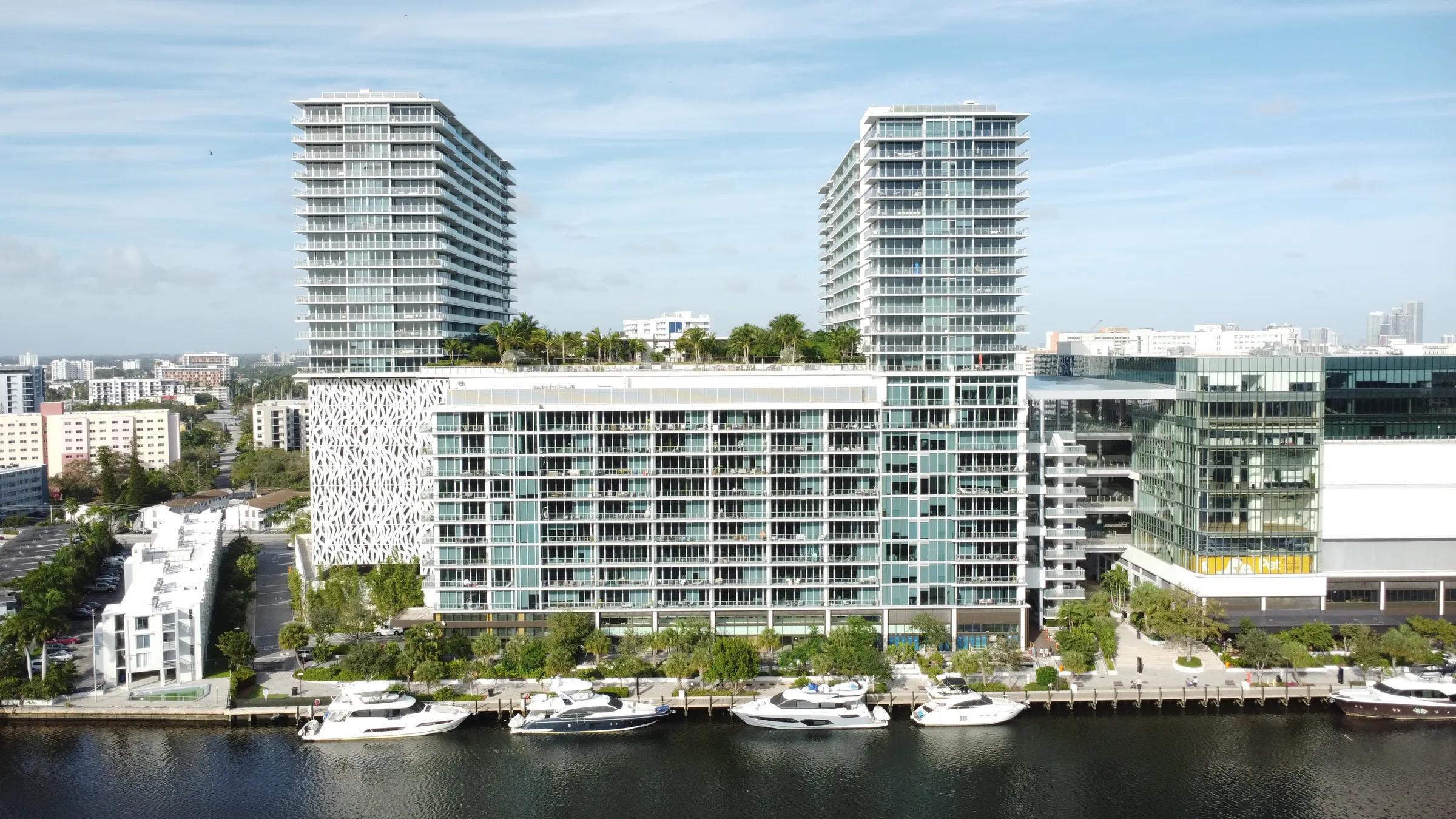 Aerial view across the Miami River of a modern condo complex with two towers, a rooftop garden and yachts docked along the riverwalk.